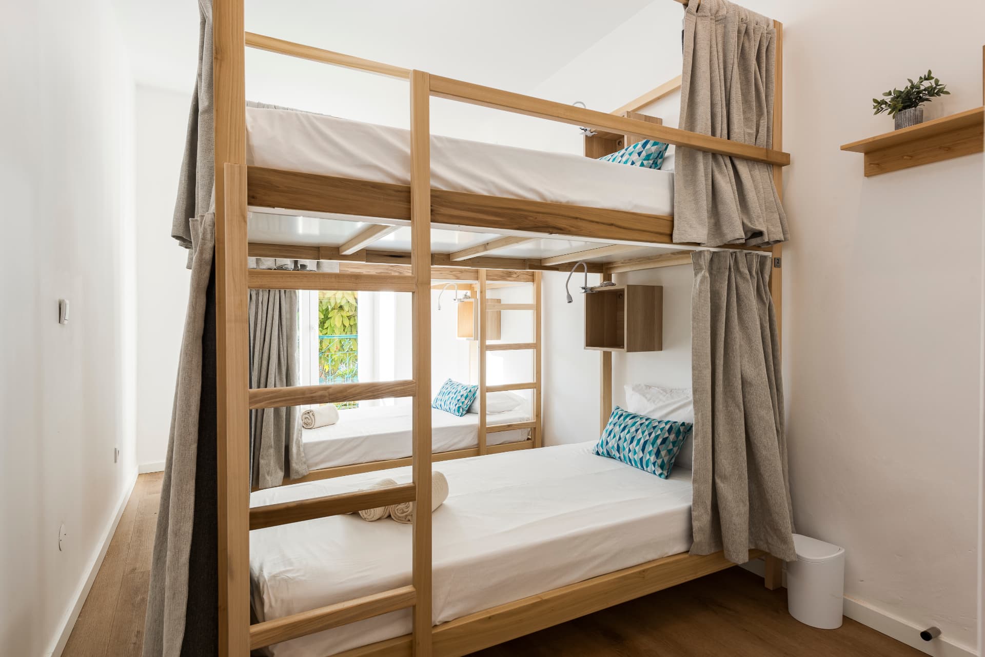 Dormitório de quatro camas do Madalena, com dois beliches de madeira, roupa de cama branca e cortinas de privacidade