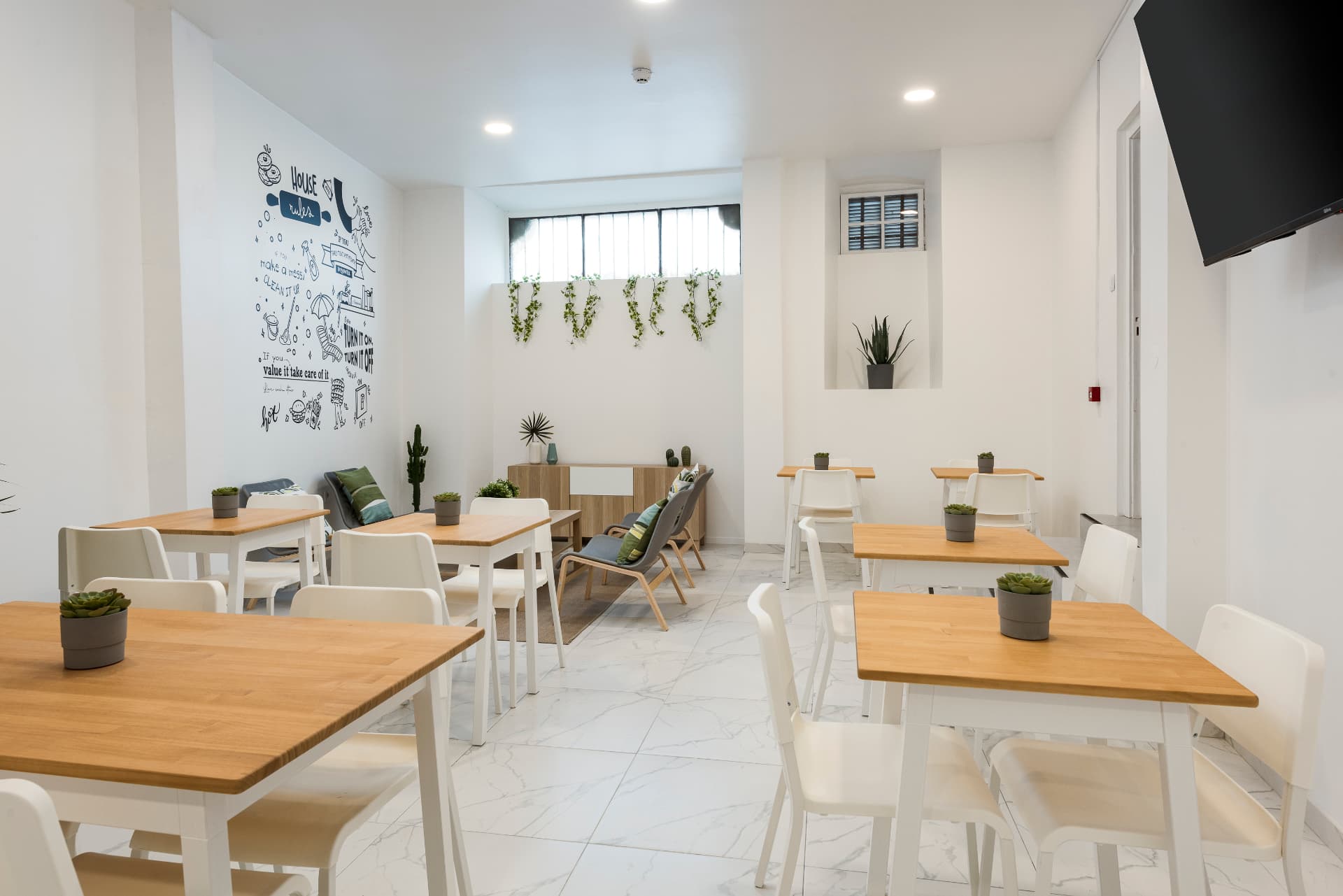 Sala de refeições e convívio do Madalena, com mesas de tampo em madeira, cadeiras brancas e mural House Rules